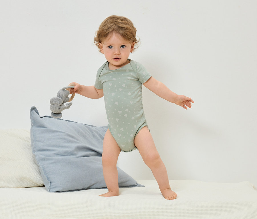Egy kisgyermek szívmintás zöld bodyban áll az ágyon, és egy szürke elefántos játékot tart a kezében.