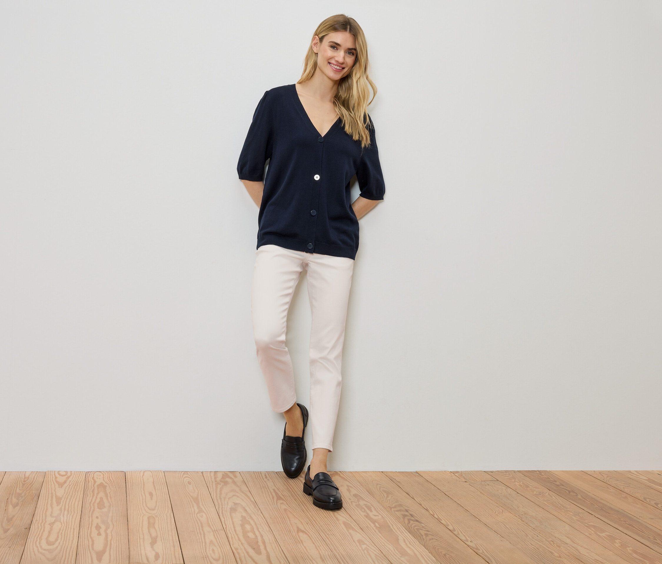 Egy nő áll a falnál, sötétkék kardigánt, világos nadrágot és fekete cipőt visel.