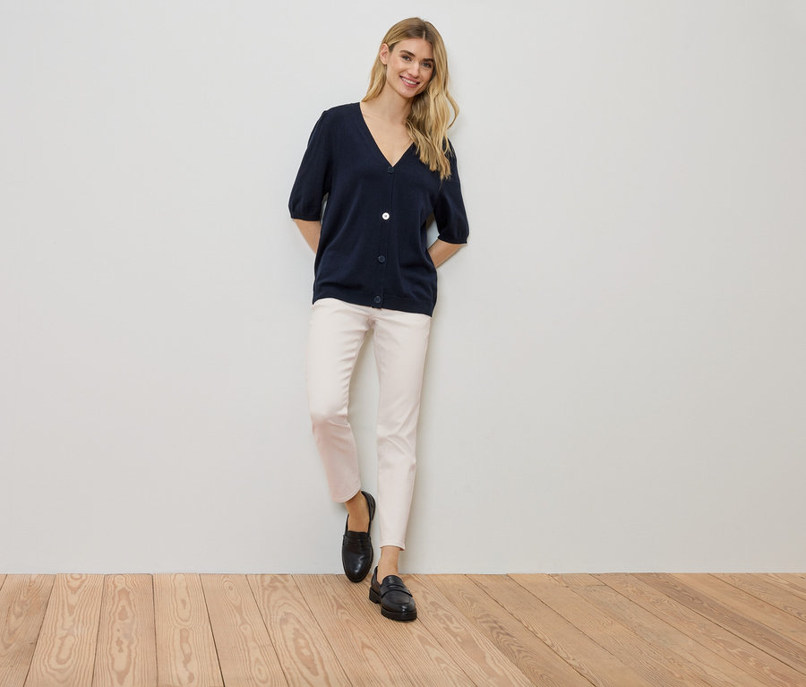 Egy nő áll a falnál, sötétkék kardigánt, világos nadrágot és fekete cipőt visel.