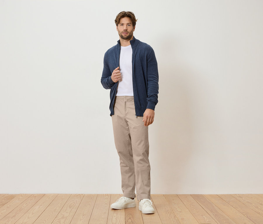 Egy férfi áll egy fehér fal előtt, sötétkék kardigánt, fehér pólót, bézs nadrágot és fehér tornacipőt visel.