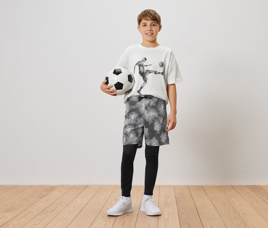 Egy fiú egy futball-labdát tart a bal kezében. Futballista mintás pólót, rövidnadrágot és leggingset, valamint fehér tornacipőt visel.