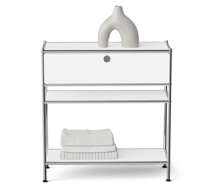 Egy fehér éjjeliszekrény króm vázzal, tetején egy szürke szoborszerű vázával és egy polcon összehajtott törölközőkkel.