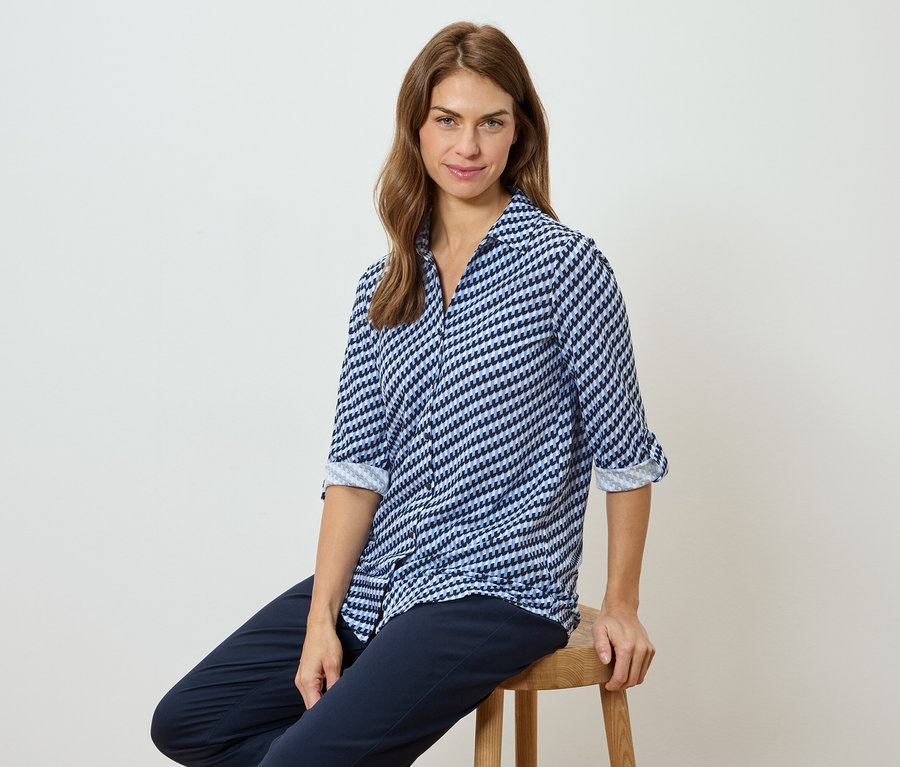 Egy nő ül egy széken, kék-fehér mintás blúzt és sötét nadrágot visel.