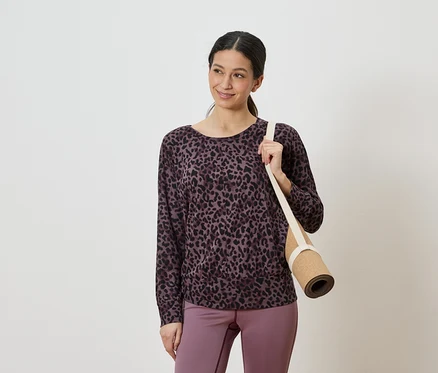 Lila-fekete leopárdmintás felsőt és lila leggingset viselő nő, aki a vállán összetekert jógamatracot visz.