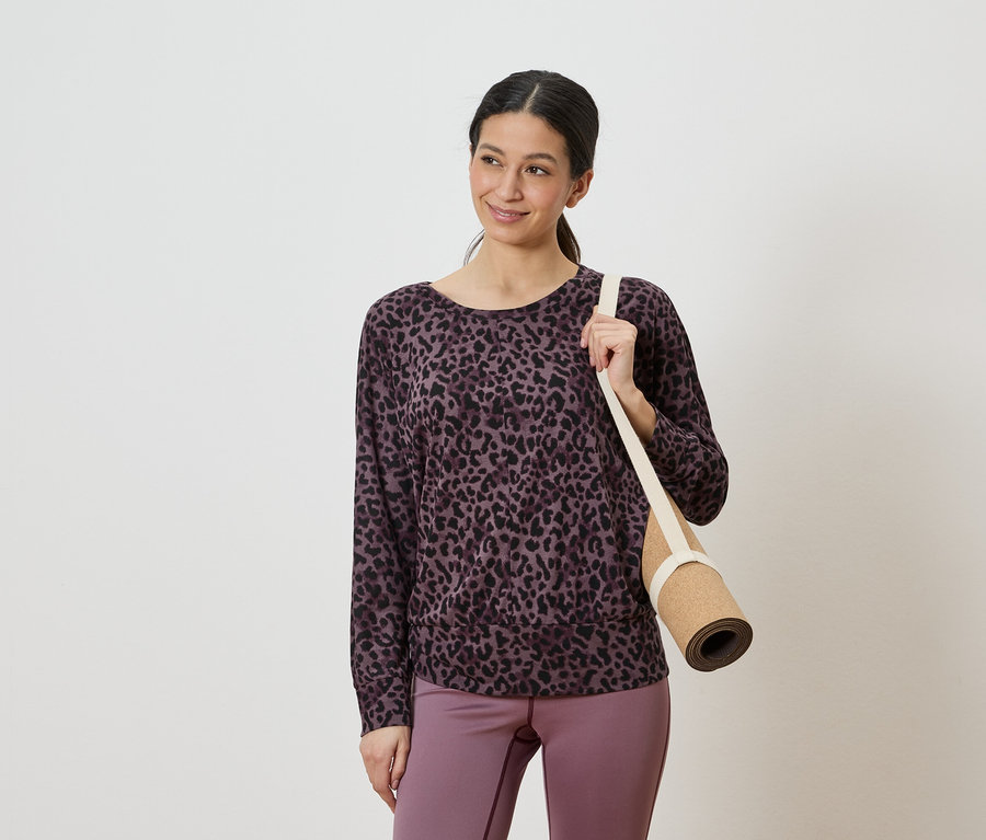 Lila-fekete leopárdmintás felsőt és lila leggingset viselő nő, aki a vállán összetekert jógamatracot visz.