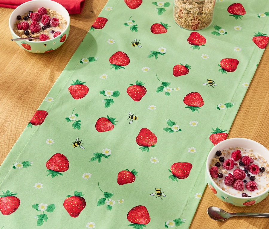 Egy fa asztalon egy epermintás asztali futó, rajta müzlis tálak, egy pohár müzli és egy üveg tej.