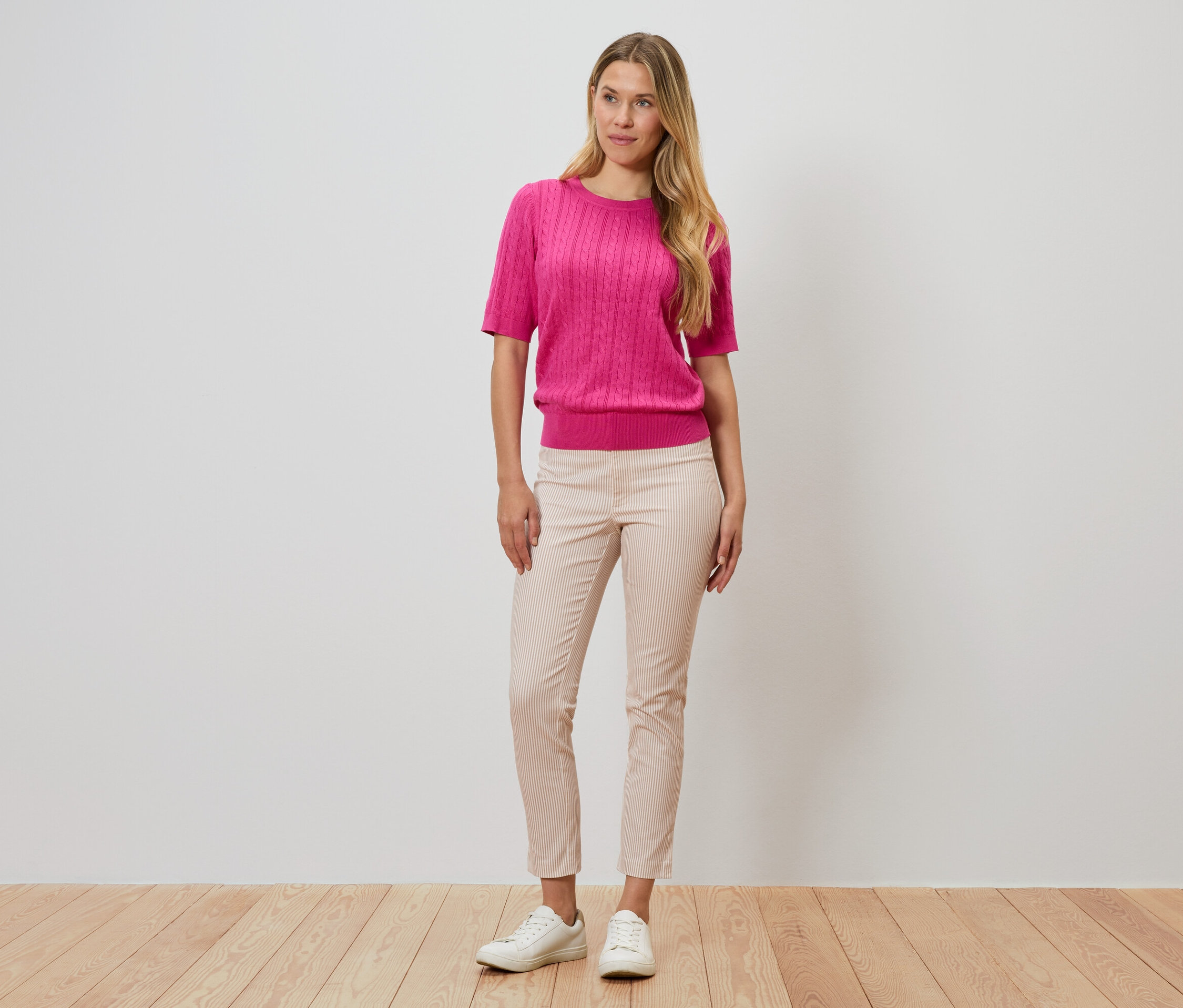 Nő áll a fal előtt, rózsaszín pulóvert, csíkos nadrágot és fehér cipőt visel.