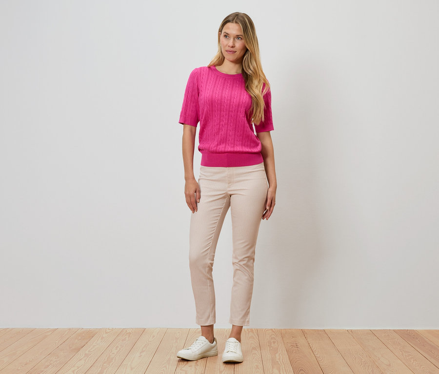 Nő áll a fal előtt, rózsaszín pulóvert, csíkos nadrágot és fehér cipőt visel.