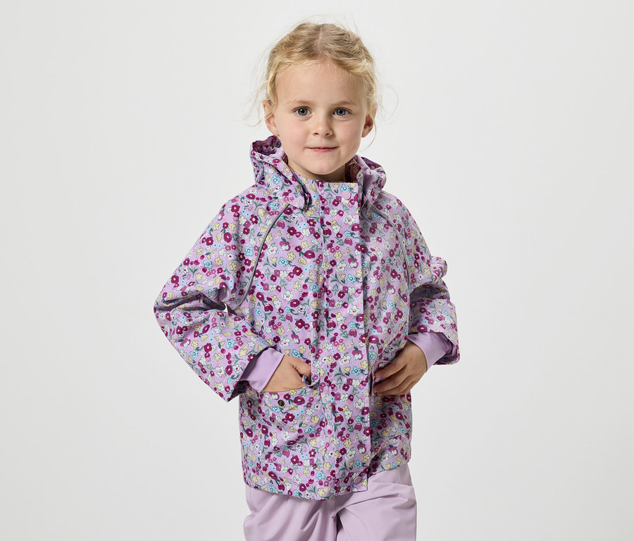 Szőke hajú lány lila virágos kabátban áll fehér háttér előtt.