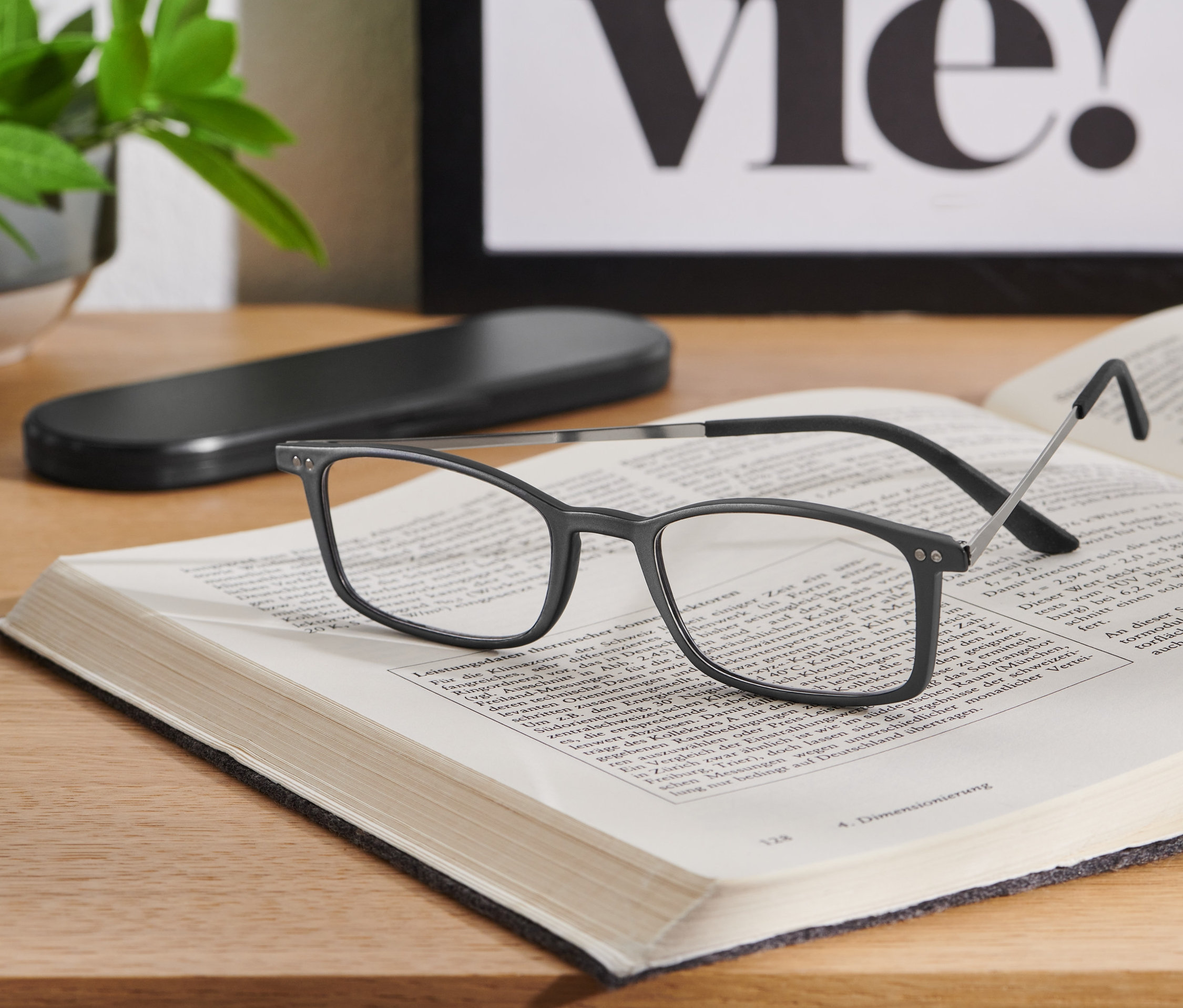 Egy fekete, ultravékony olvasószemüveg fekszik egy nyitott könyvön egy fa asztalon.