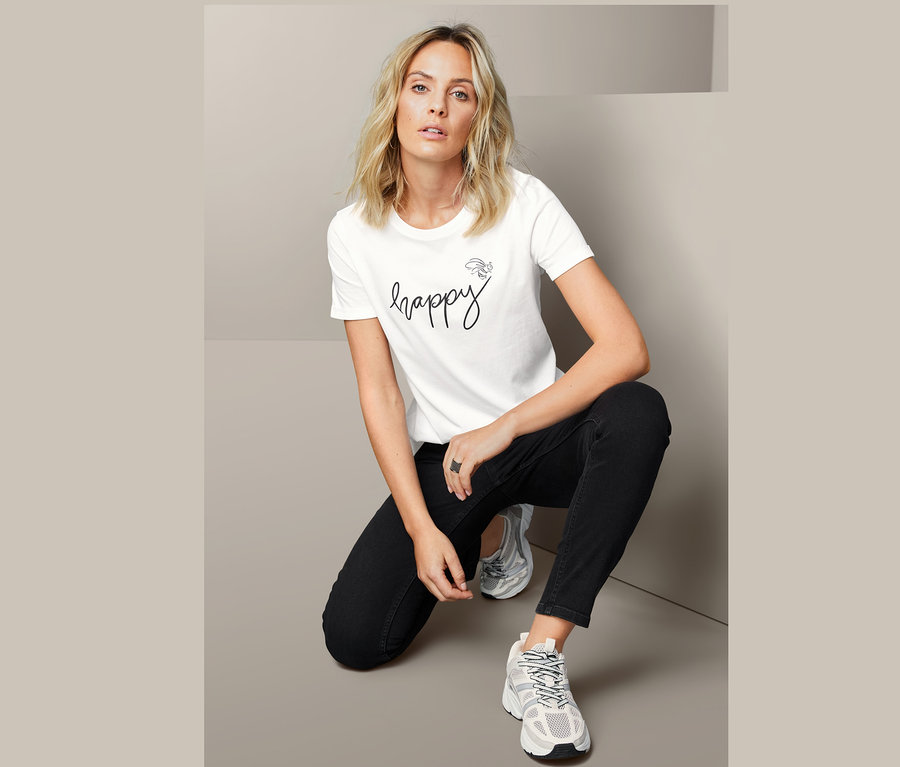 Nő térdel, és egy fehér, "happy" feliratú női pólót visel.