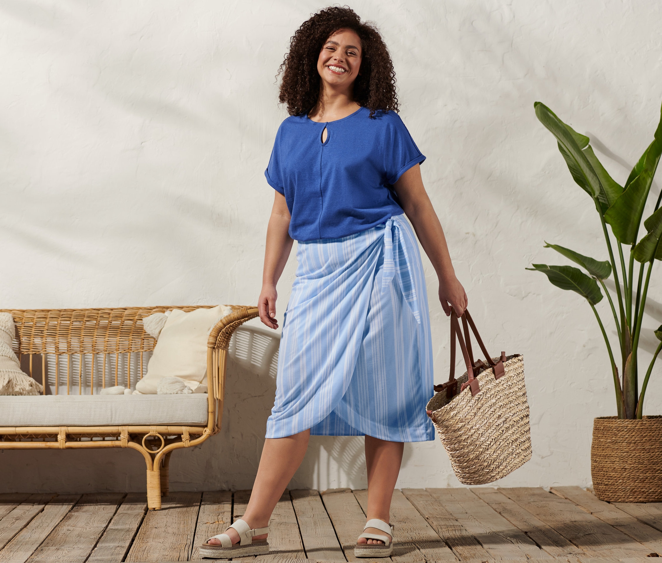 Nő visel egy lenvászon anyagú felsőt, egy csíkos megkötős szoknyát, egy bőr szandált és egy kosártáskát.