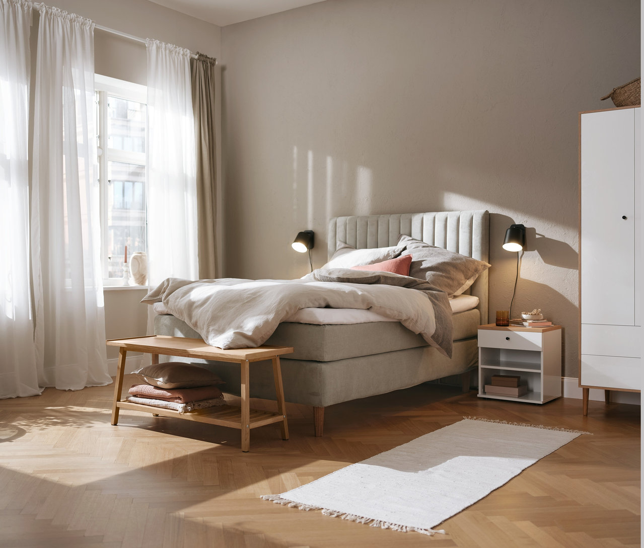 Bézs boxspring ágy (kb. 180 x 200 cm), Bekeson ruhásszekrény és éjjeliszekrény, fekete fém fali lámpa és ülőpad cipőtartóval.