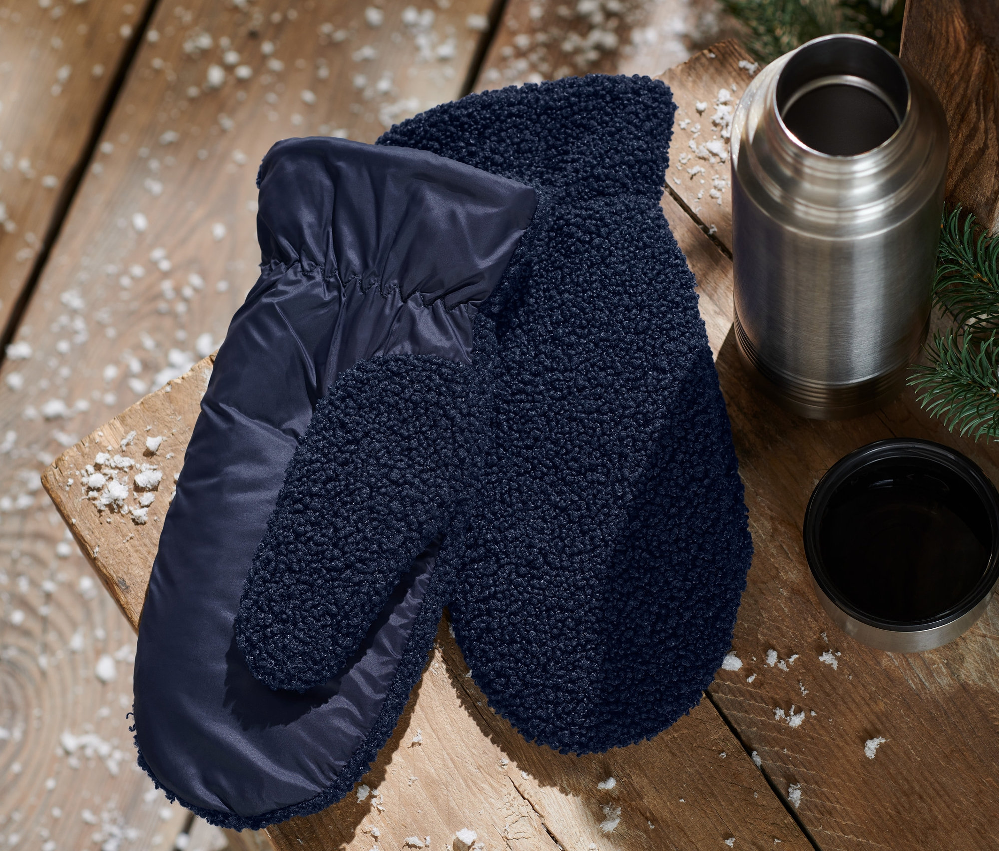 Egy fa asztalon kék női egyujjas kesztyű, plüss, sötétkék, egy termosz és egy csésze található.
