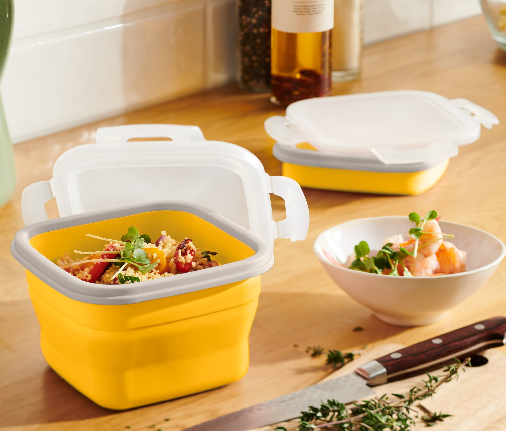 Egy sárga, összehajtható ételhordó doboz tartalmmal egy fa asztalon áll egy zöld kancsó és egy garnélarákos tál mellett.