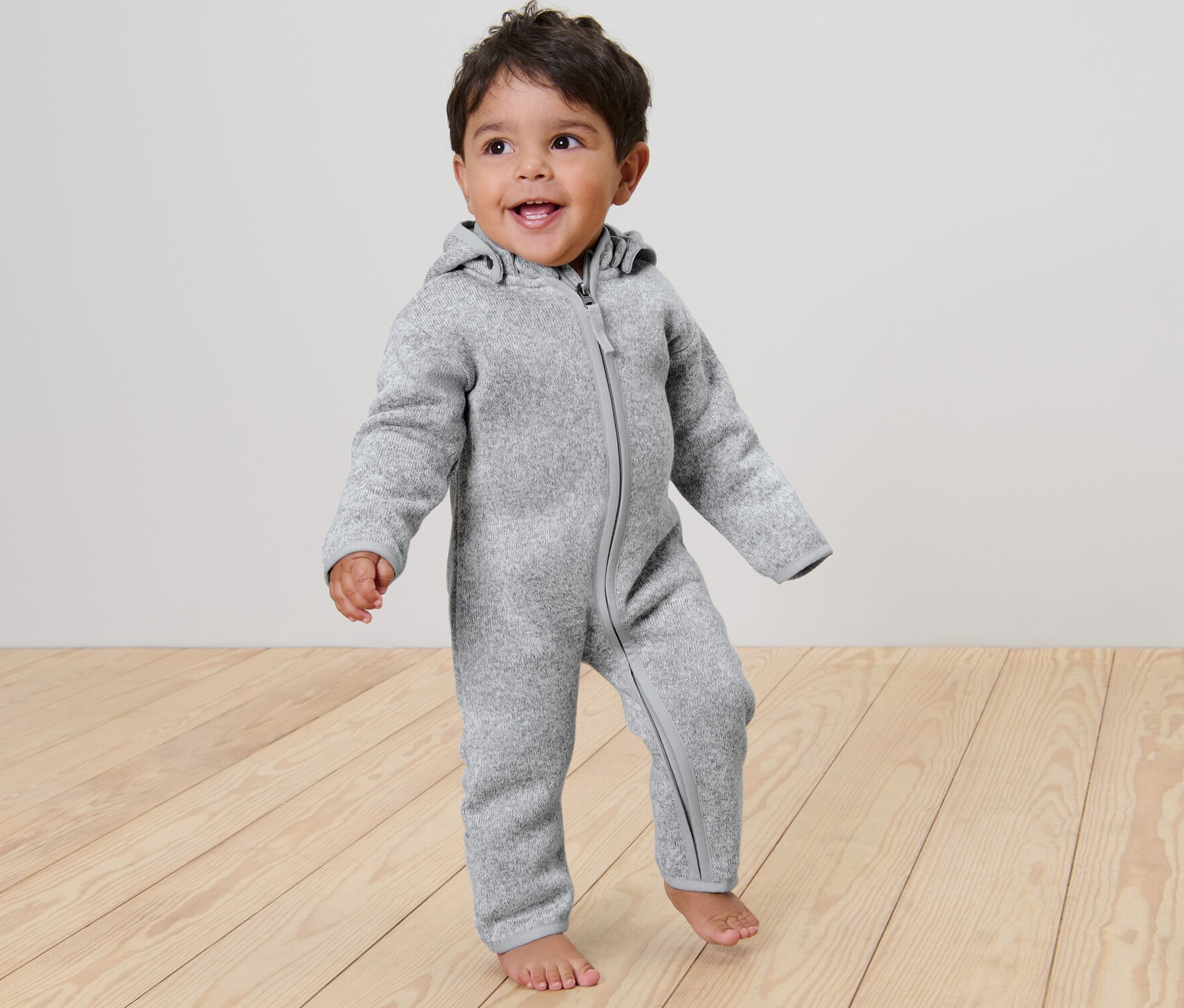 Egy mosolygó gyermek áll egy fapadlón, szürke kötött polár kezeslábast visel.