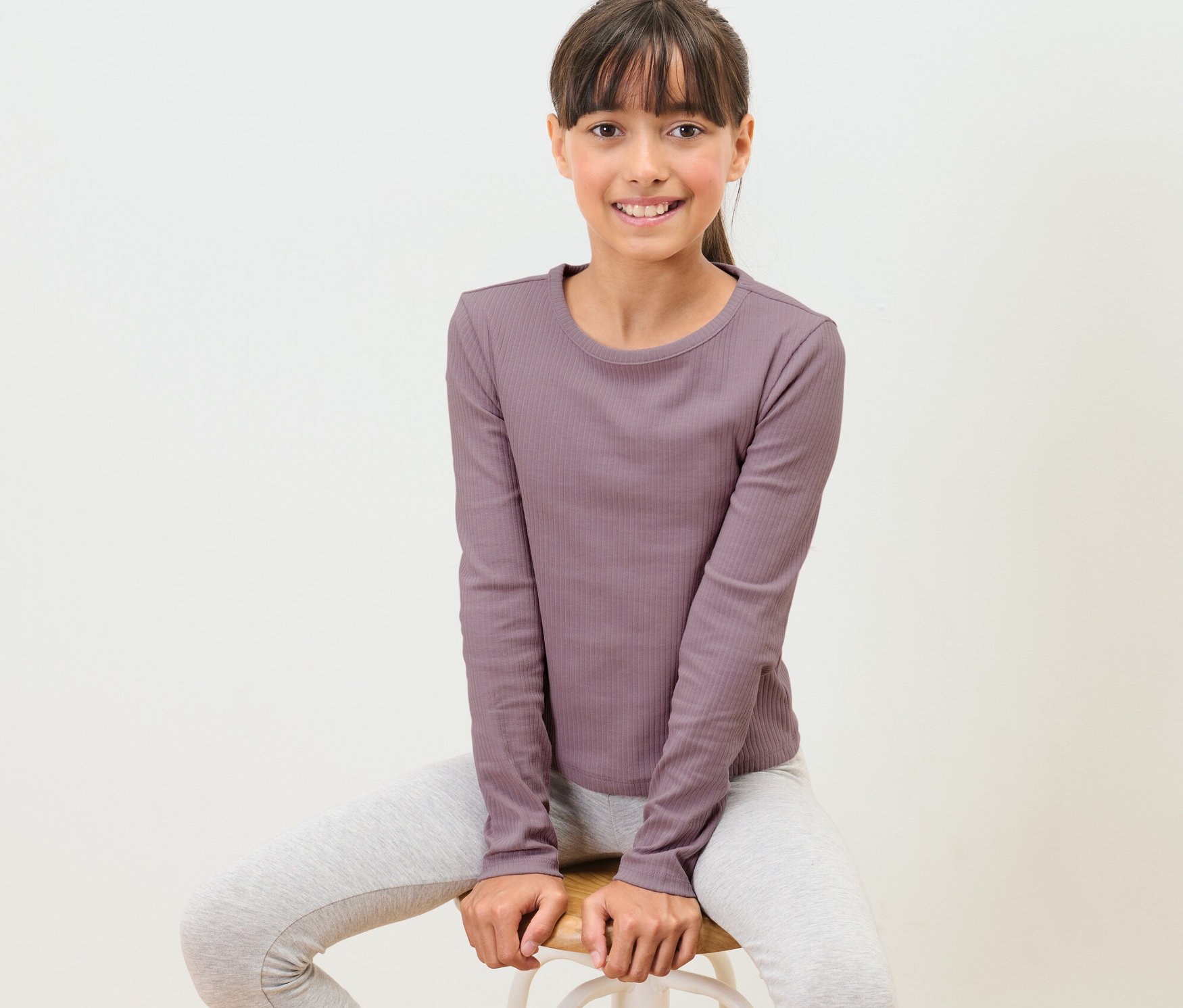 Egy lány ül egy széken, és mályva színű, bordázott hosszú ujjú felsőt visel.