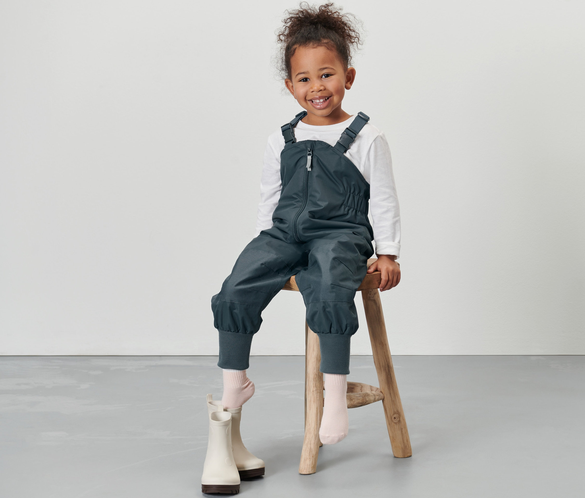 Mosolygó kislány ül a sámlin, pala kék kisgyerek termo esőnadrágot visel.