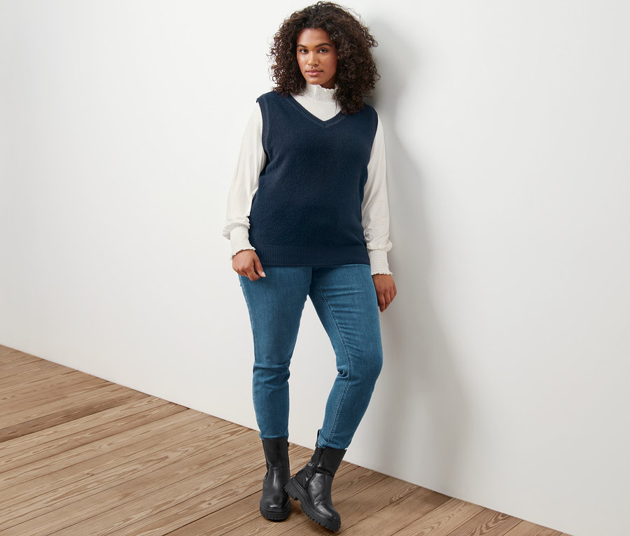 Nő pózol buklé hatású kötött mellényben, krém színű állógalléros ingblúzban és farmer hatású Fit Nelly treggingsben.