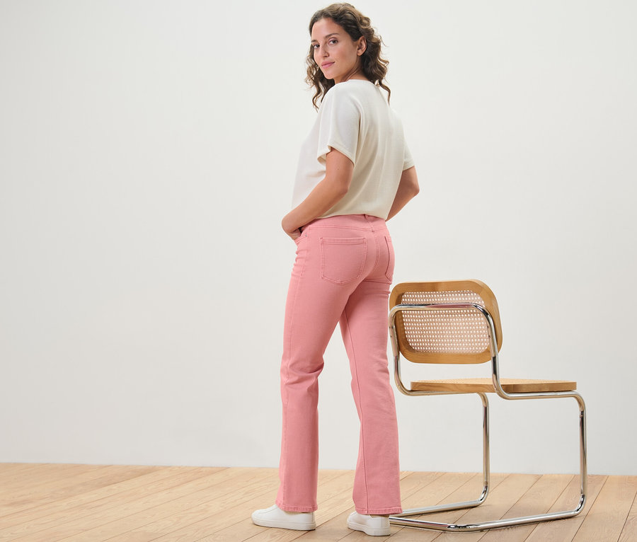 Nő pózol korall színű, »Lou« szabású bootcut farmernadrágban egy szék mellett.
