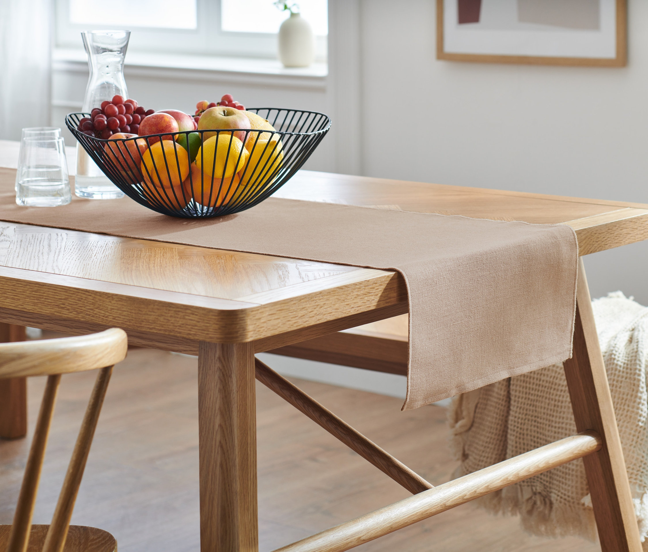 A fa asztalon egy bézs színű asztali futó található. Rajta egy drótkosár gyümölcsökkel.