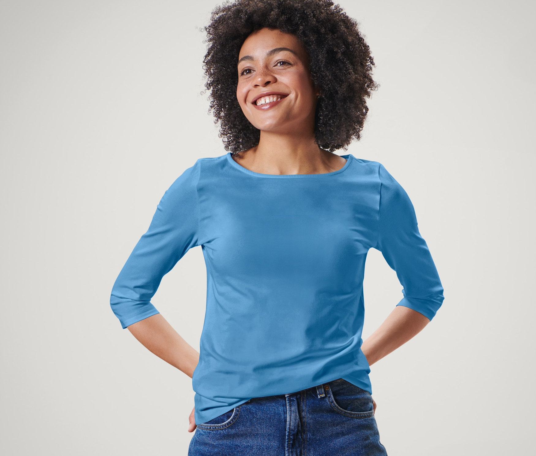 Nő pózol világoskék, 3/4-es ujjú felsőben és kék farmerben.