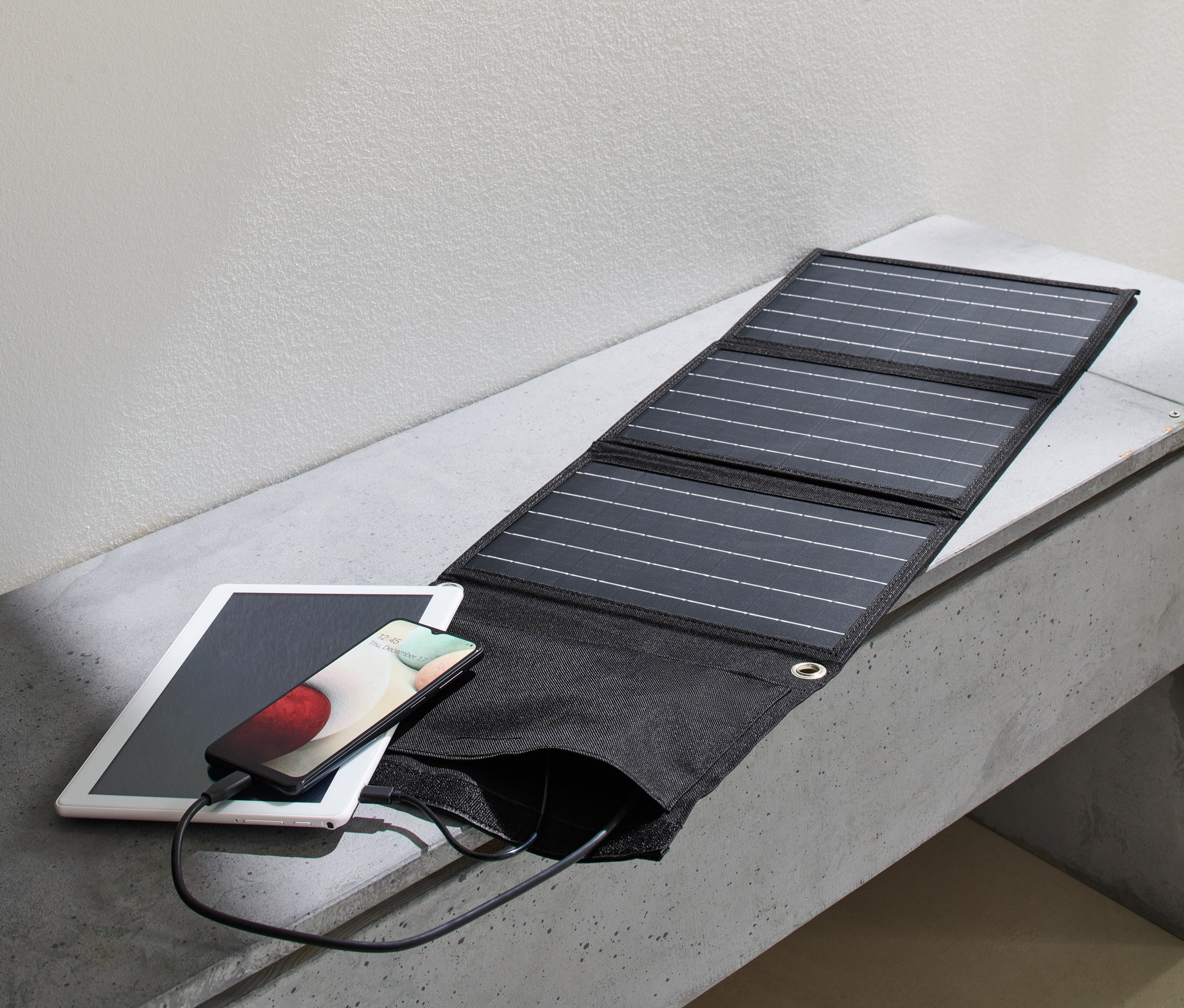 Egy összehajtható napelemes töltő tölt egy telefont, ami egy tableten fekszik egy betonpadon. Mellette egy hátizsák.