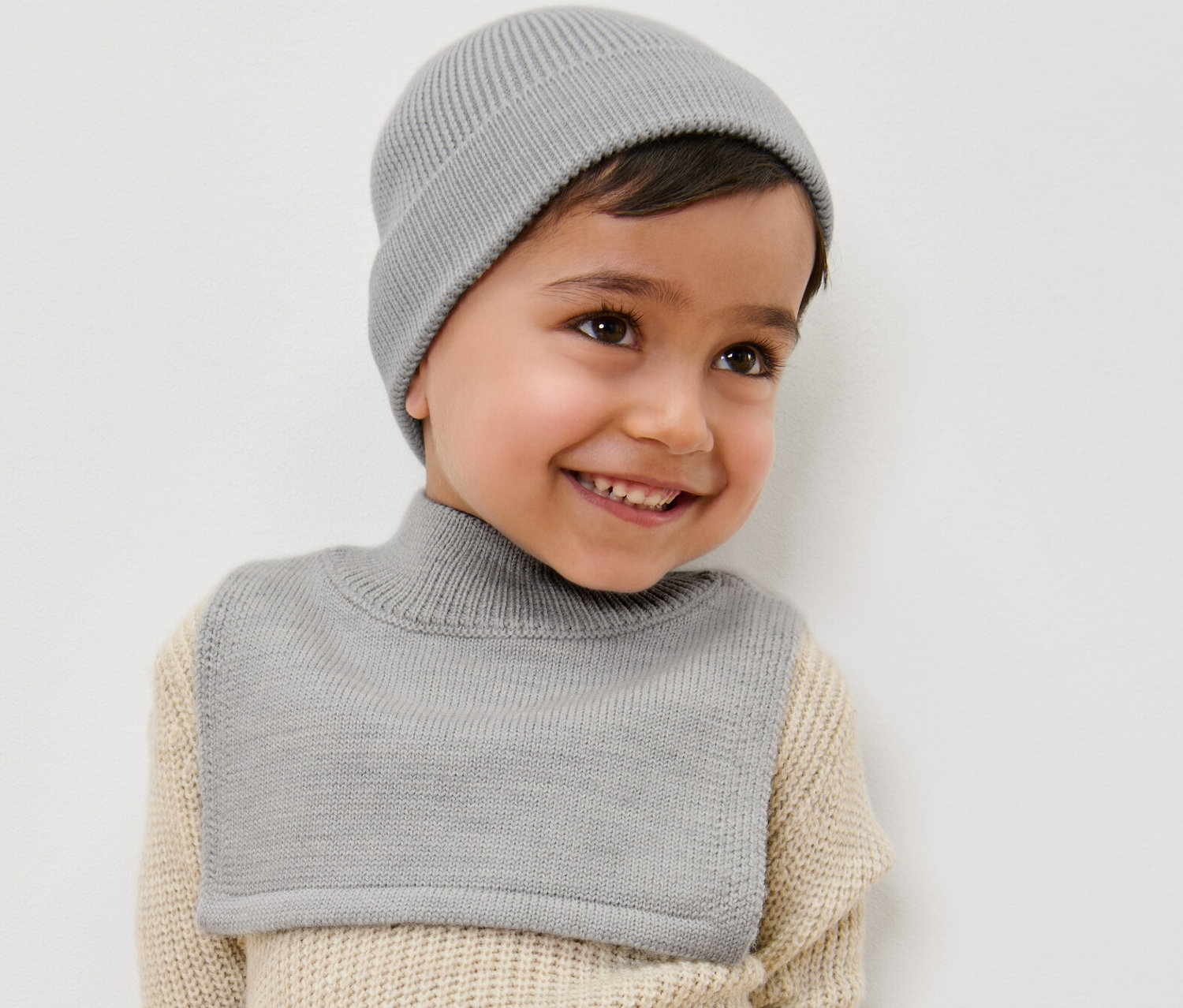Egy mosolygó kisfiú szürke gyapjú garbósálat és sapkát visel.