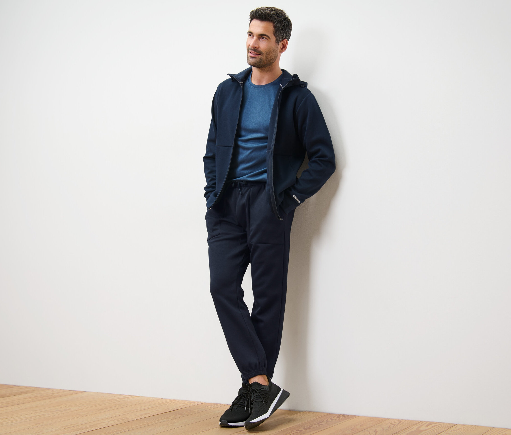 Férfi támaszkodik a falnak, sötétkék kapucnis pulóvert, pólót és férfi funkcionális szabadidőnadrágot visel.
