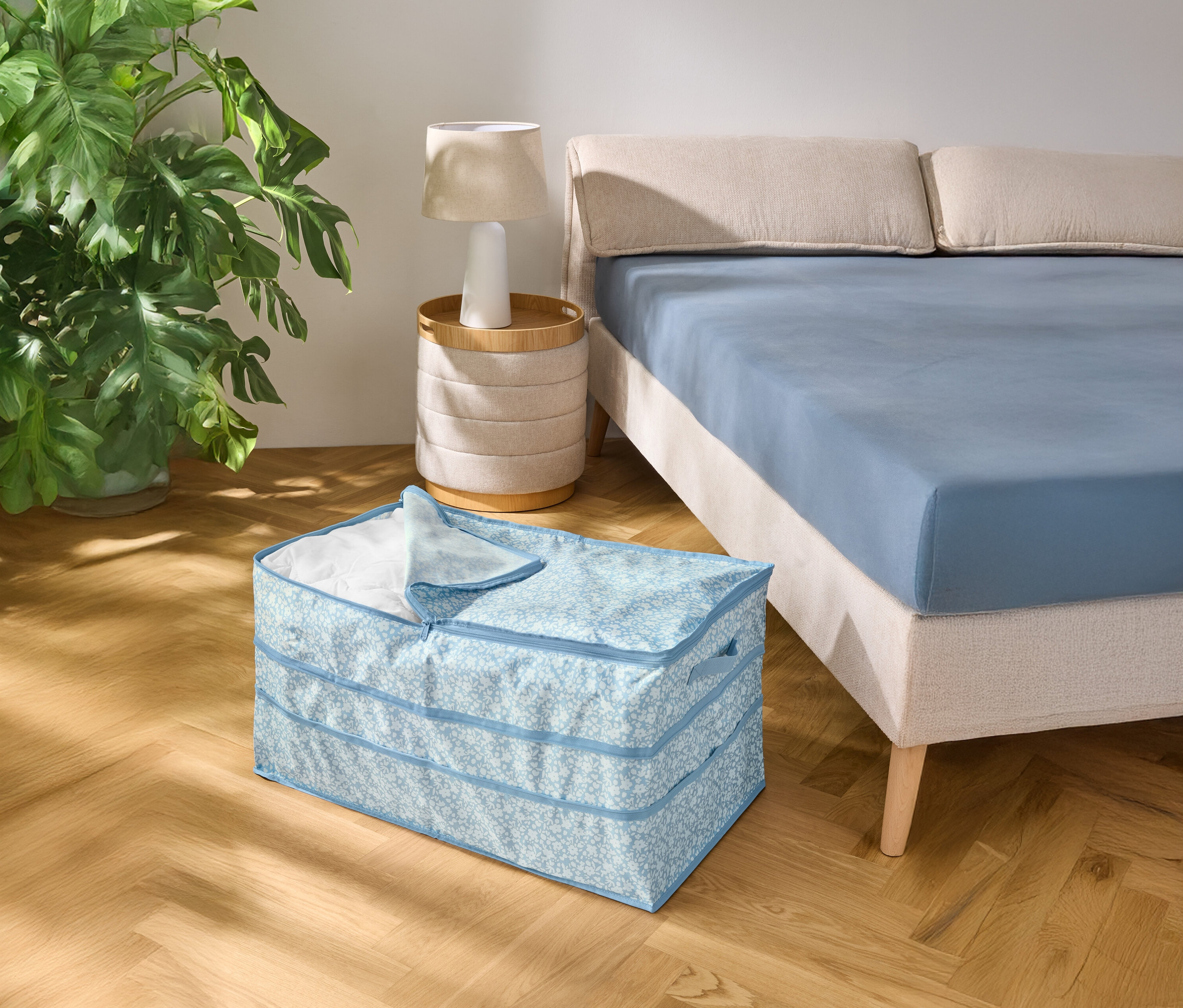 A fa padlón egy világoskék mintás tárolódoboz áll egy kék lepedővel bevont ágy mellett.