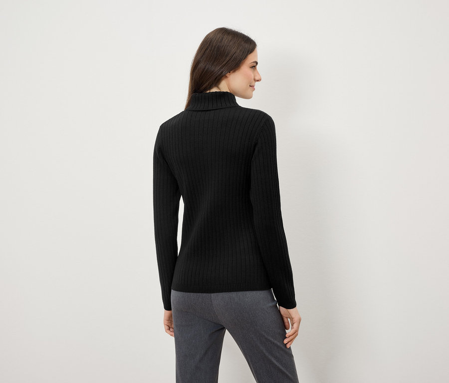 Nő fekete garbóban és szürke nadrágban áll háttal a kamerának.