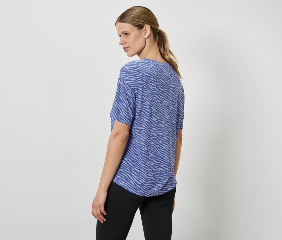 Egy nő kék, állatmintás felsőben és fekete nadrágban a válla fölött néz.