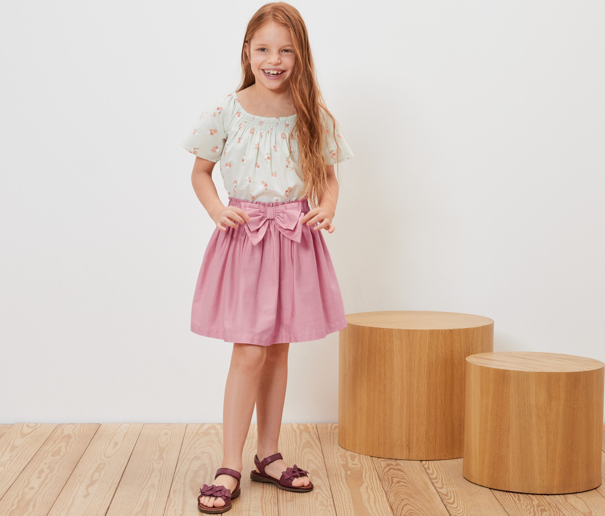Mosolygó, vörös hajú lány pózol szövött gyerekszoknyában és virágos felsőben.
