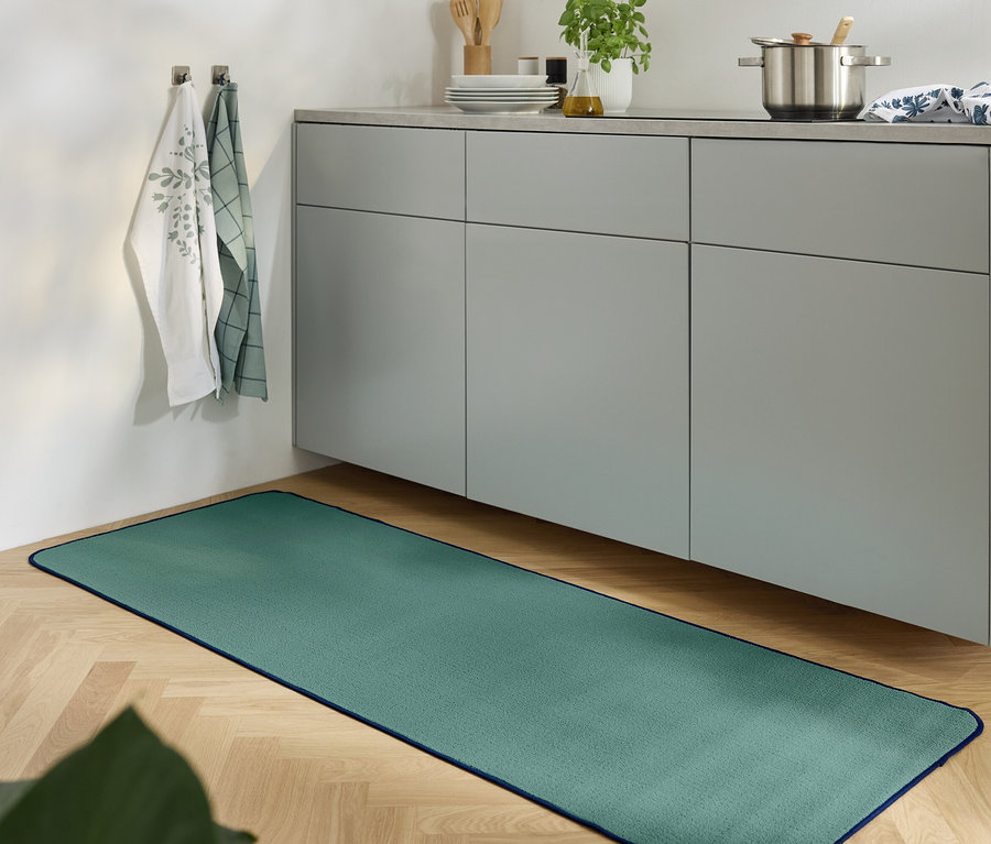 Egy zöld szőnyeg fekszik a fapadlón egy konyhában, szürke szekrénnyel.