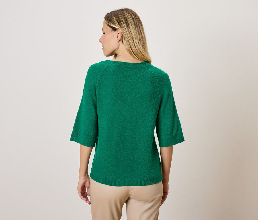 Egy nő zöld, háromnegyedes ujjú pulóvert és bézs nadrágot visel, háttal a kamerának.