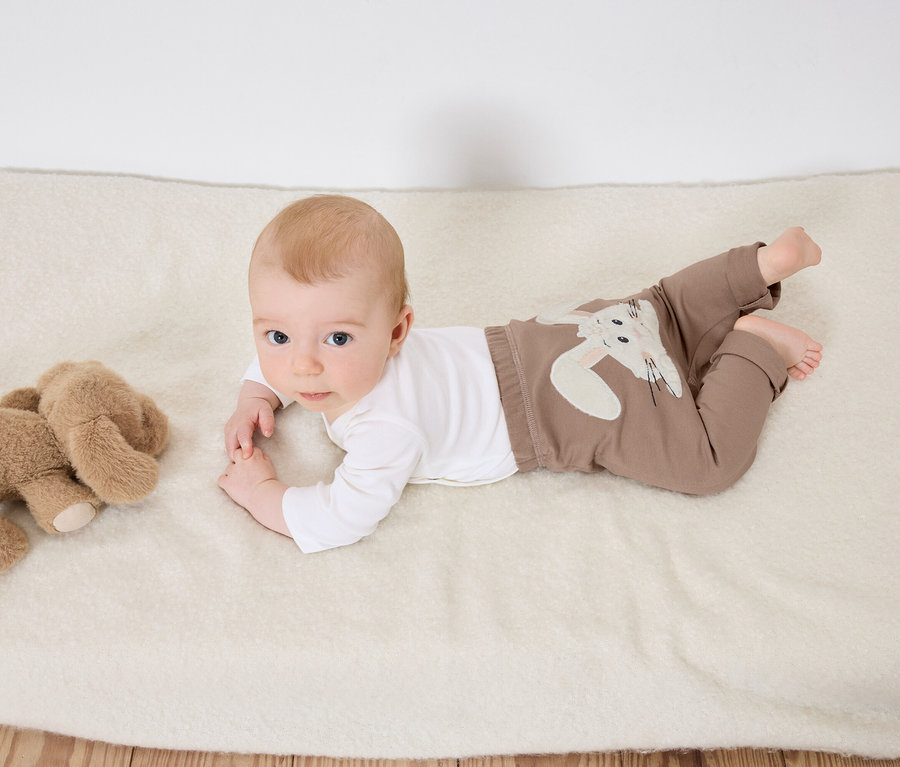 Egy baba hason fekszik egy fehér takarón egy plüss elefánt mellett.