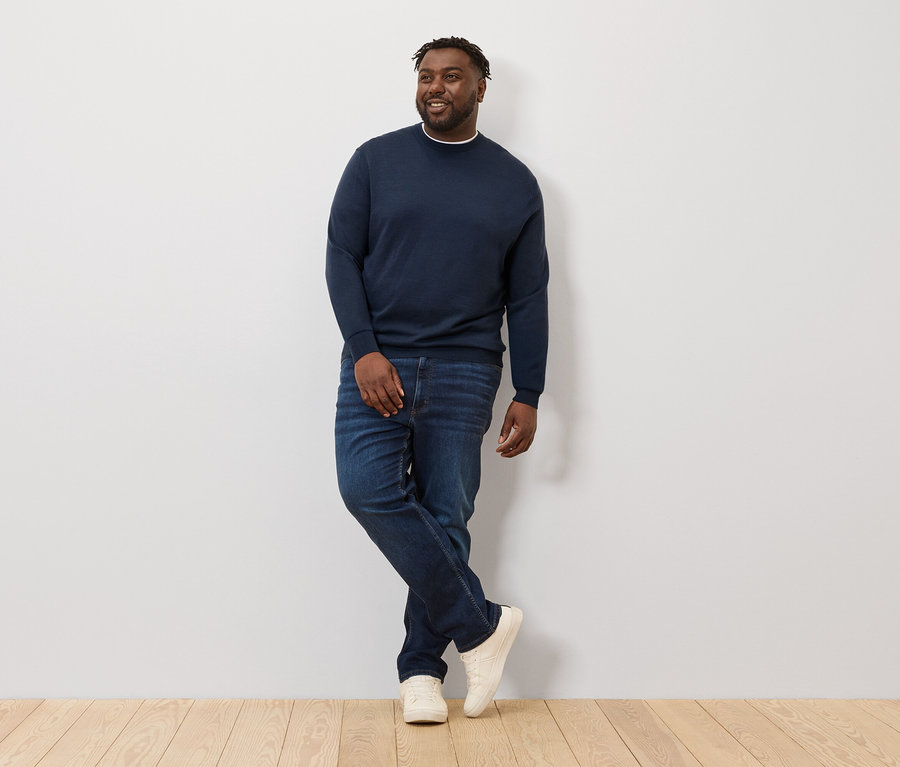 Férfi pózol a falnak dőlve, sötétkék színű, melírozott merinó gyapjú pulóvert visel.