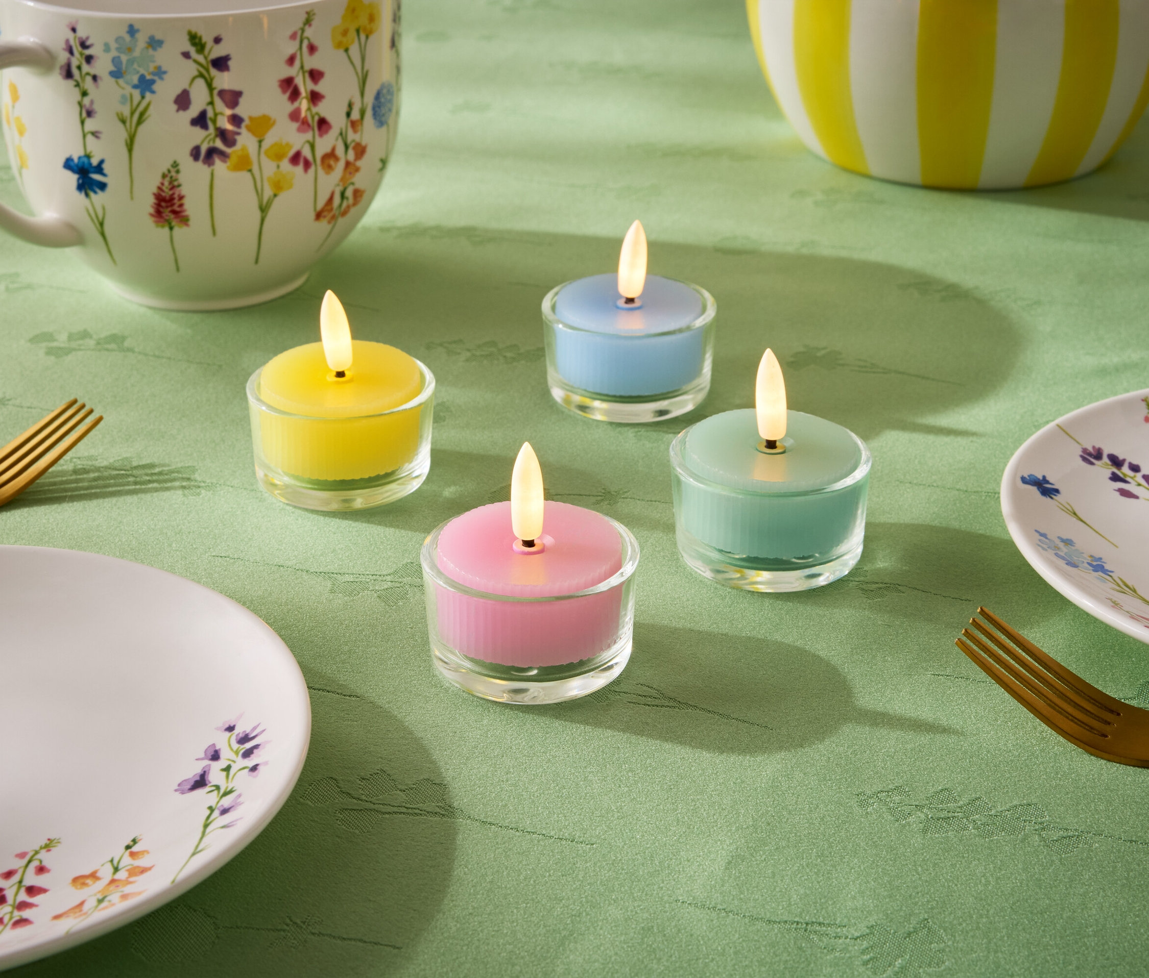 A zöld terítőn négy kis gyertya áll pasztell színekben: sárga, kék, zöld és rózsaszín. Emellett egy tányér, egy csésze és egy virágmintás tál, valamint két arany villa is látható.