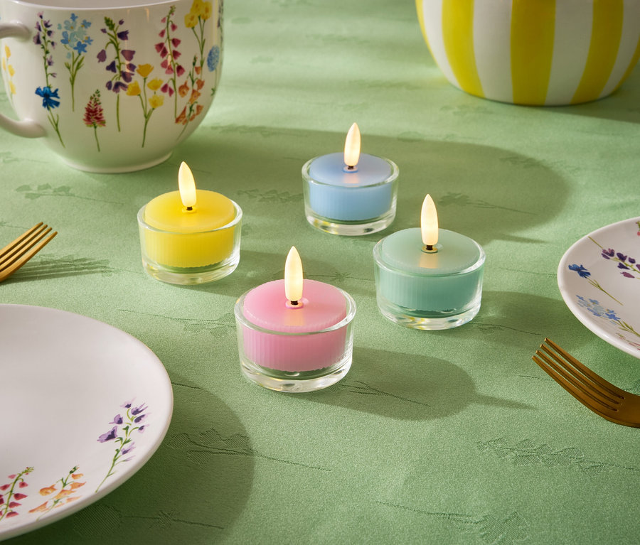 A zöld terítőn négy kis gyertya áll pasztell színekben: sárga, kék, zöld és rózsaszín. Emellett egy tányér, egy csésze és egy virágmintás tál, valamint két arany villa is látható.