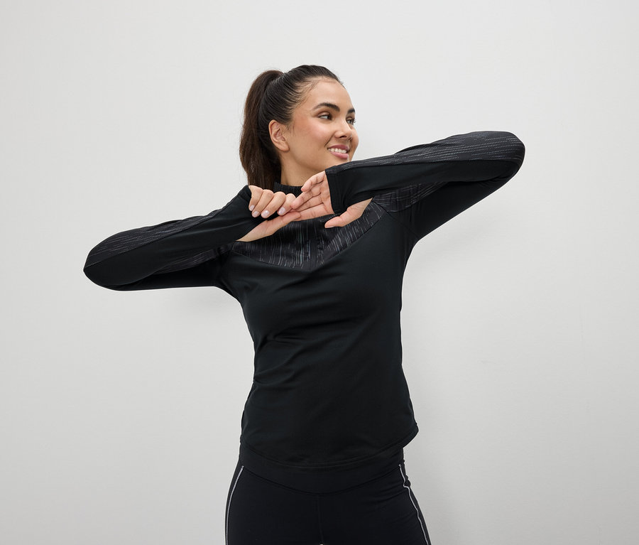 Mosolygó nő fekete sportruházatban nyújtózkodik fehér háttér előtt.