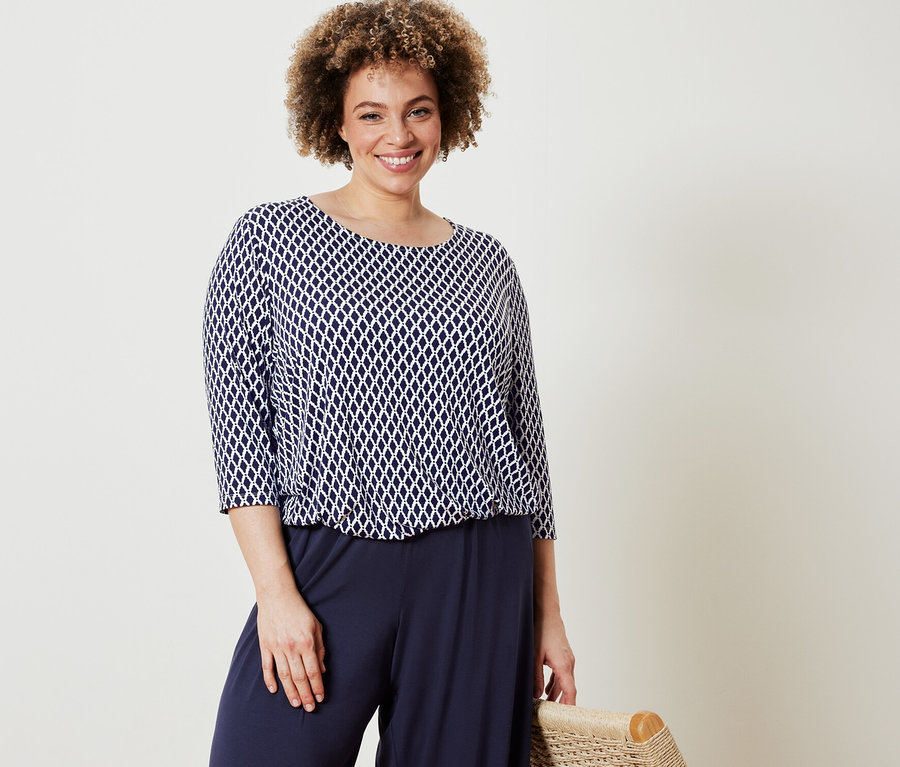 Nő pózol mintás pólóblúzban és sötétkék, bő szárú nadrágban.