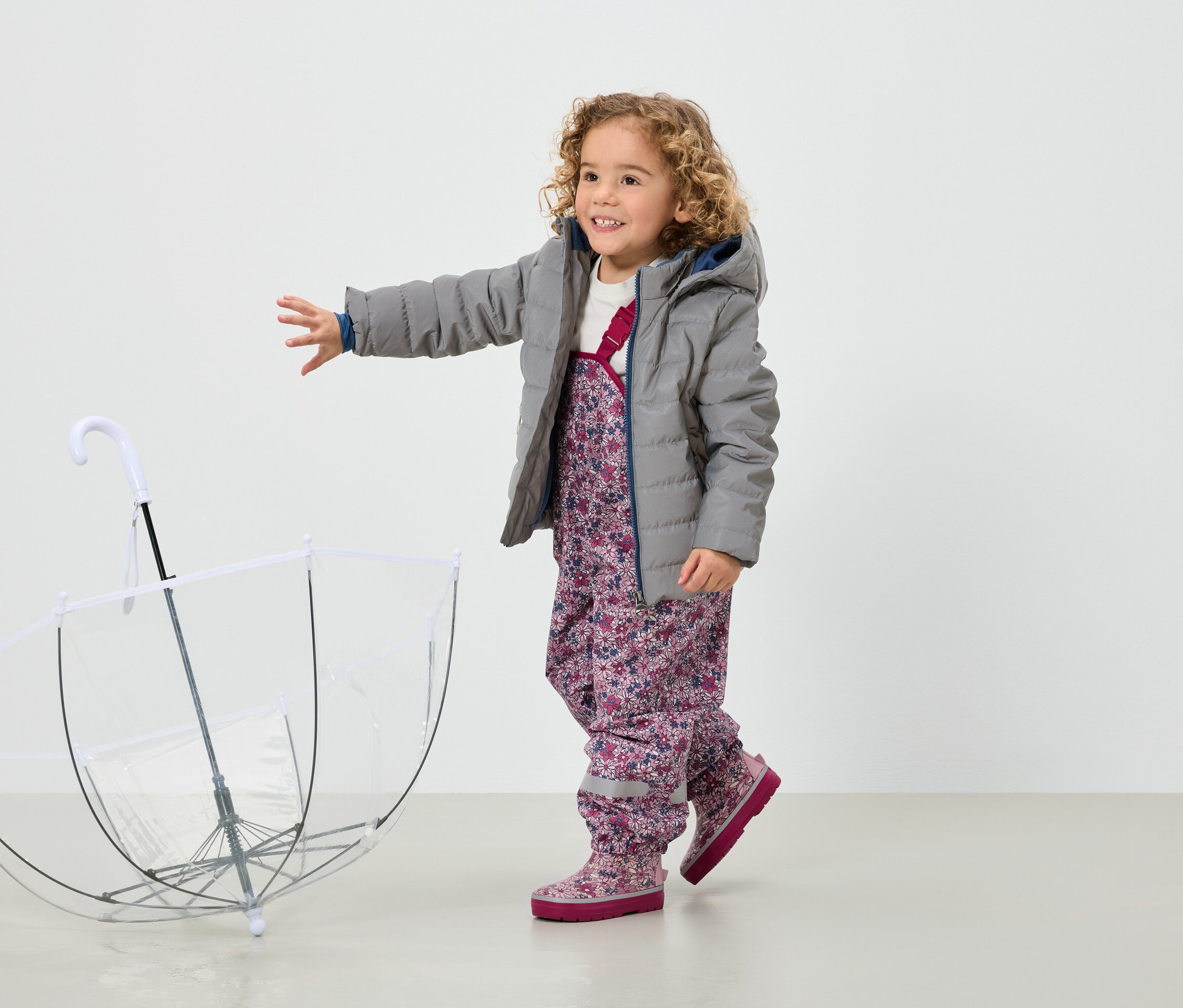 Egy mosolygós, göndör hajú kislány szürke kabátban és virágos nadrágban áll egy átlátszó esernyő mellett.