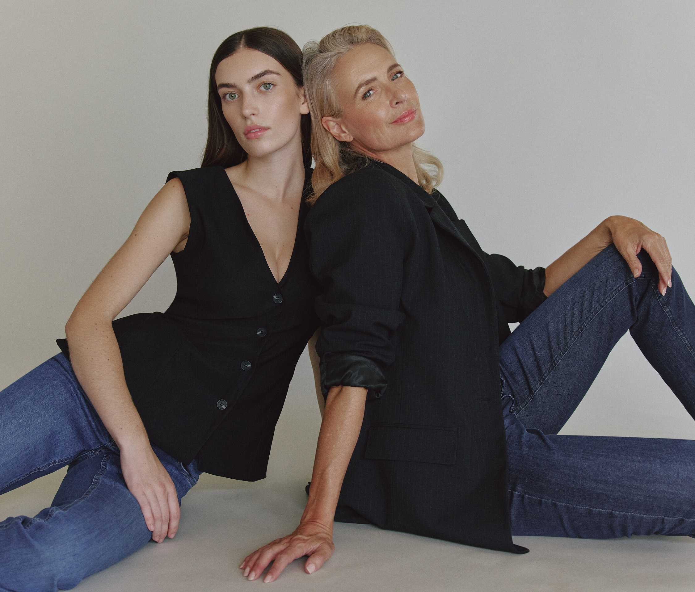Két nő ül egymásnak háttal a földön. Az egyik fekete zakót és farmert, a másik fekete mellényt és farmert visel.