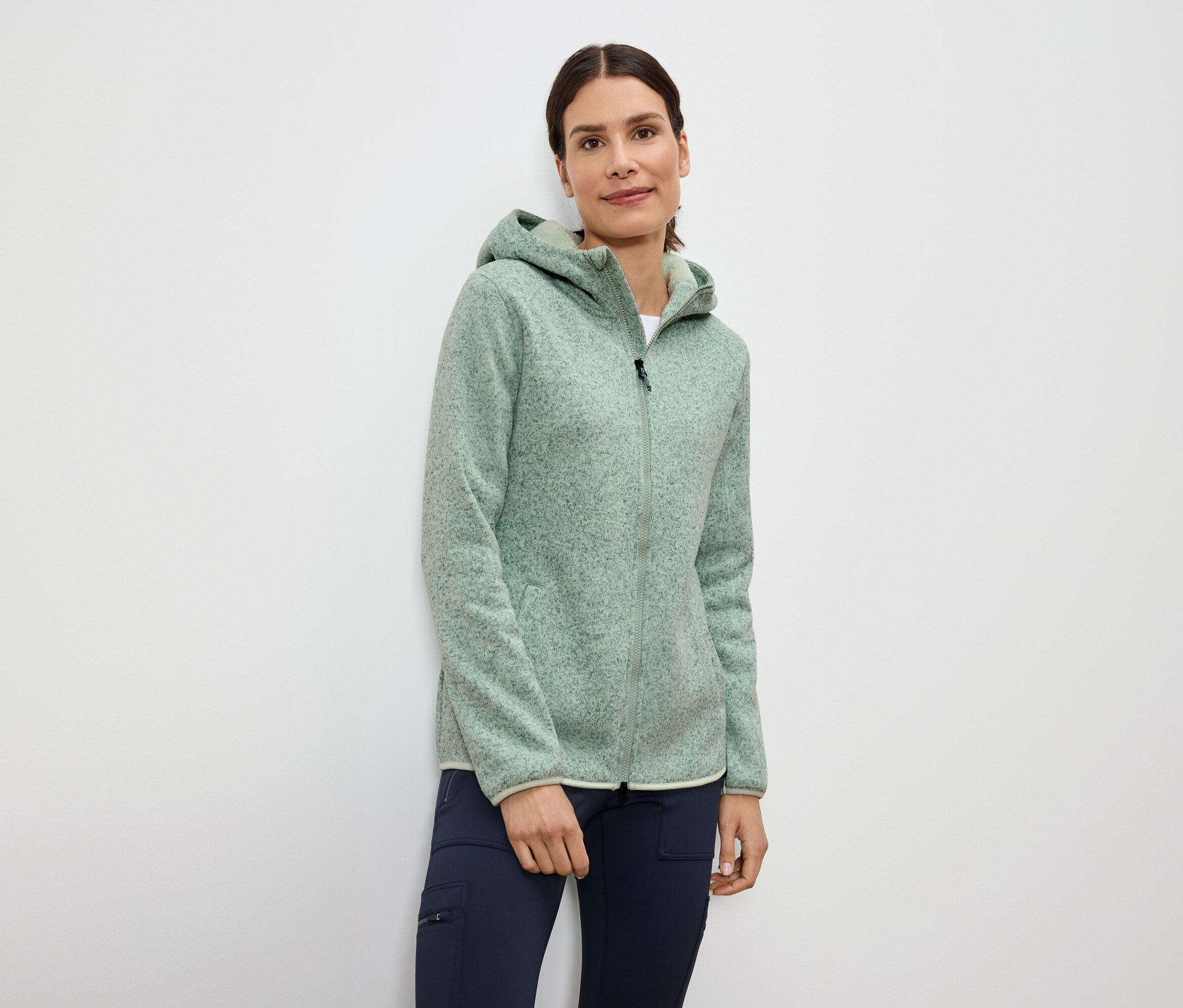 Nő visel világoszöld, cipzáras, kapucnis polár pulóvert és sötétkék nadrágot.