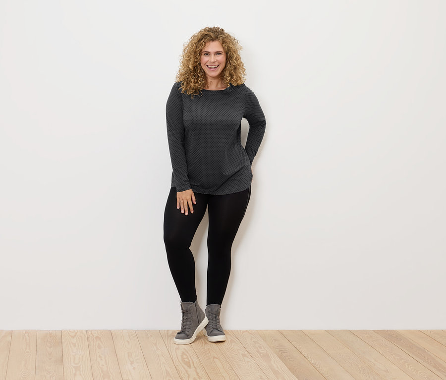 Göndör hajú nő áll és mosolyog, fekete leggingset, szürke cipőt és szürke-fekete pólót visel.