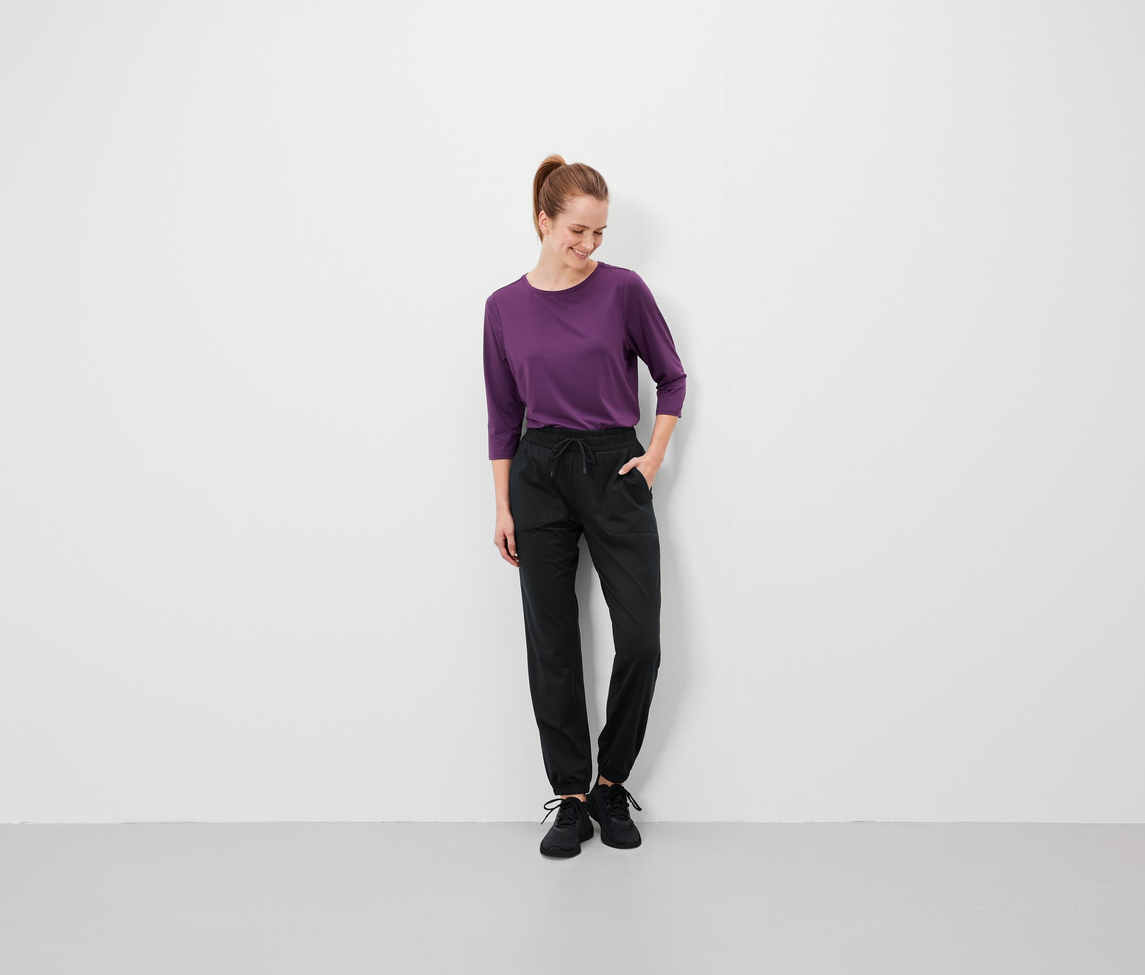 Barna hajú nő lófarokkal áll, kezét a fekete nadrágja zsebébe teszi, és lila pólót visel.