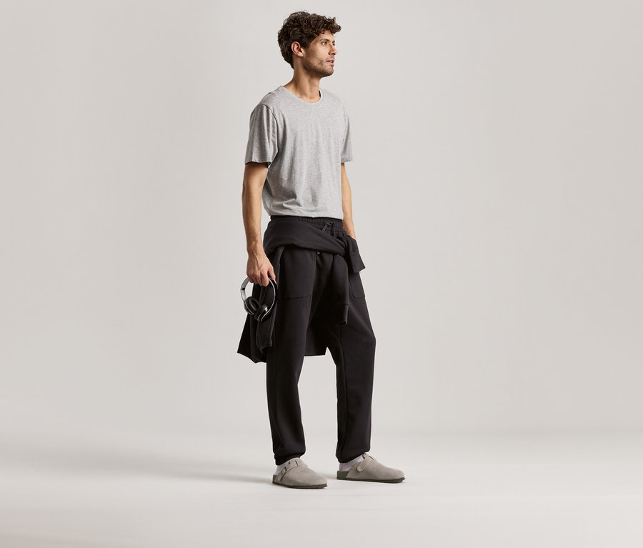 Férfi szürke pólót és fekete szabadidőnadrágot visel. A kezében fejhallgatót tart.