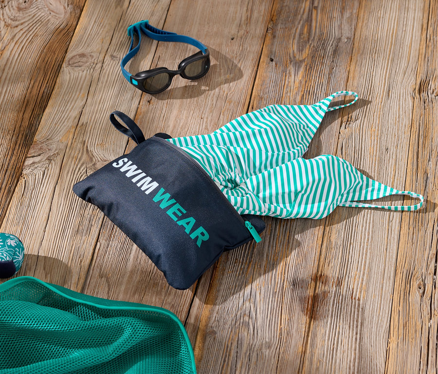 Fotó úszási kiegészítőkről egy fa felületen, beleértve egy „Swimwear” feliratú táskát, egy zöld-fehér csíkos fürdőruhát, egy úszószemüveget és egy zöld hálót.