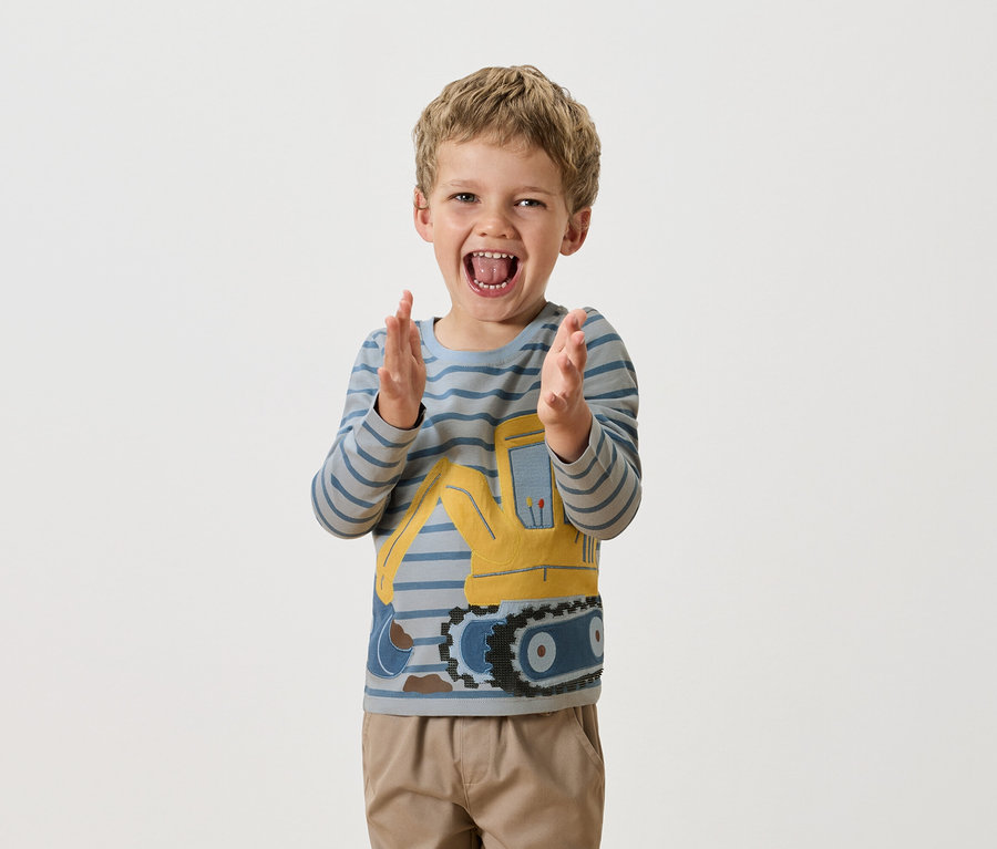 Egy szőke hajú fiú tapsol, kék csíkos, hosszú ujjú pólót visel, amin egy sárga buldózer van.