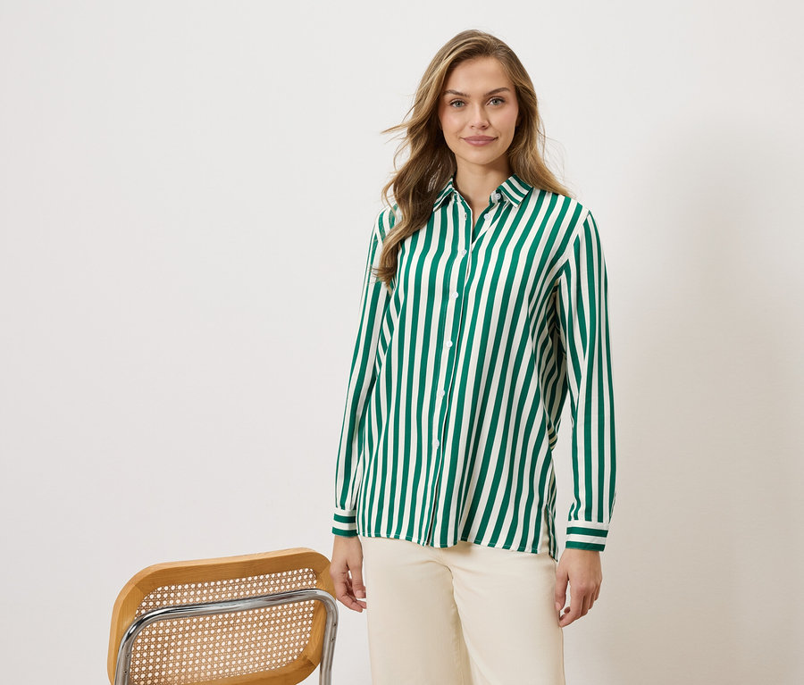 Nő visel egy zöld-fehér csíkos inget és bézs nadrágot, egy szék mellett.